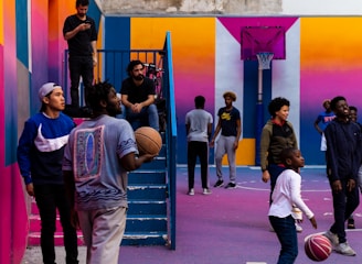 A vibrant outdoor basketball court filled with people of various ages engaged in different activities. Some are playing basketball while others are standing and chatting. The court is surrounded by brightly painted walls in shades of pink, orange, and blue. A basketball hoop is mounted on the back wall, adding to the sports theme. A few people are sitting on stairs or using phones, creating a relaxed and social atmosphere.