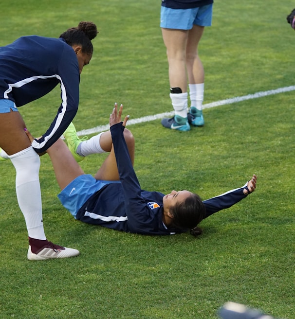 A group of athletes in sportswear are on a green soccer field. Two individuals are interacting closely; one helps the other who is lying on the grass. Nearby, other athletes are stretching.