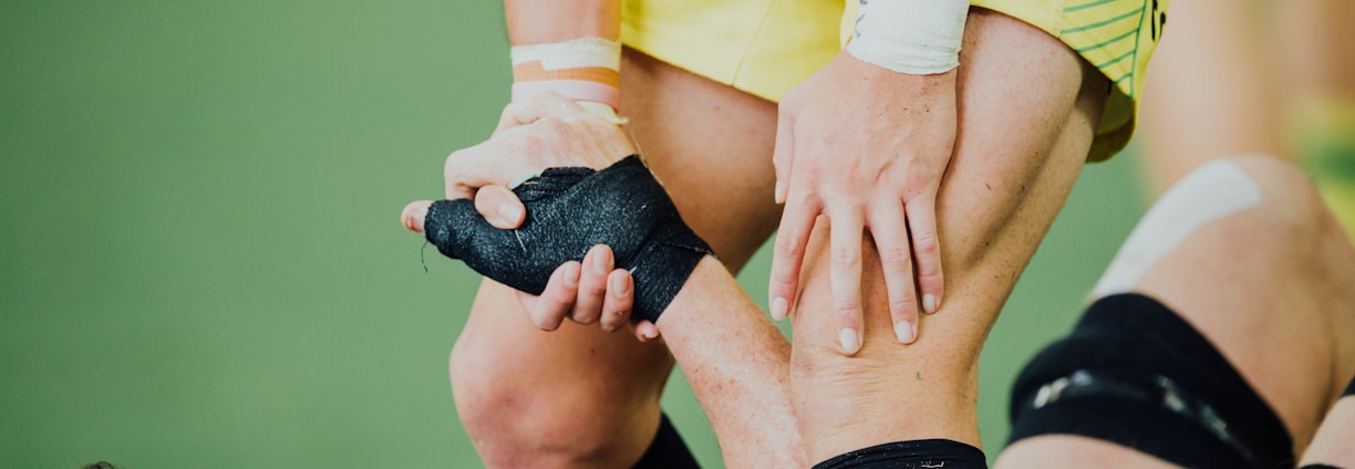 A close-up of a sports interaction where one person in a yellow uniform helps another wearing black leg gear. The person in yellow is gripping the leg of the other individual, providing support. The background appears to be a green sports field.