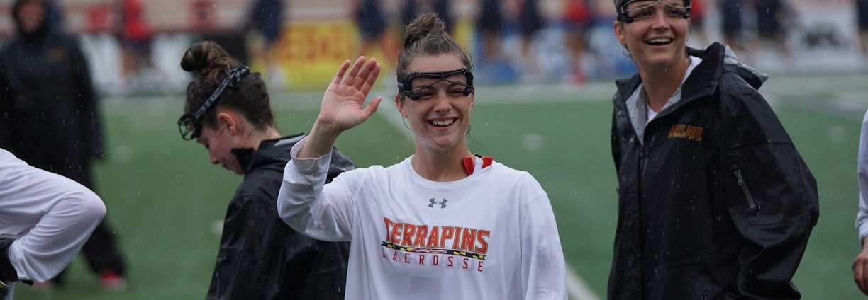 Several athletes in sports attire are on an outdoor sports field, interacting and smiling. One individual in the foreground waves with a friendly expression, while another stands beside her, also smiling. They wear protective goggles and gear associated with lacrosse. The background features indistinct figures in dark clothing against a backdrop of stadium seating.