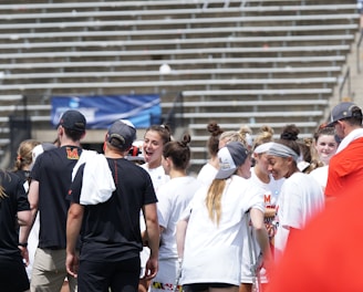 A group of athletes in white shirts with black and red accents are gathered together, engaged in conversation and smiling. They appear to be on a sports field with empty bleachers in the background. The setting is outdoors, and it seems sunny. Some individuals are wearing caps or headbands.