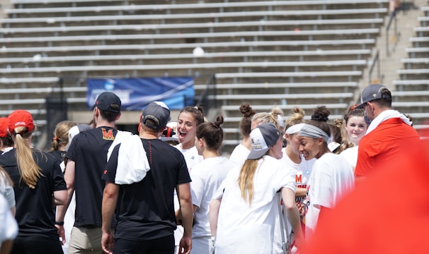 A group of athletes in white shirts with black and red accents are gathered together, engaged in conversation and smiling. They appear to be on a sports field with empty bleachers in the background. The setting is outdoors, and it seems sunny. Some individuals are wearing caps or headbands.