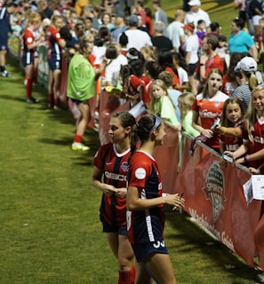The image features a lively gathering of young soccer players and fans next to a field. Several female athletes in red and blue uniforms interact with an enthusiastic crowd standing behind a barrier. Some people in the crowd are holding objects, like pens and paper, possibly for autographs.