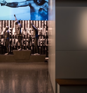 A dimly lit display features statues of athletes standing on a podium. They are positioned in front of a backdrop with the words 'LEVELING THE PLAYING FIELD' in bold letters. On the wall to the right, a softly lit quote is partially visible. The overall ambiance suggests a museum or exhibition setting.