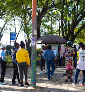 A group of people gathers in a park under leafy trees. They are interacting near a kiosk or stand surrounded by greenery and paved ground. Some individuals are wearing colorful clothing, and one person is wearing a number 7 sports jersey. There's a mixture of standing and walking activities happening in the scene.