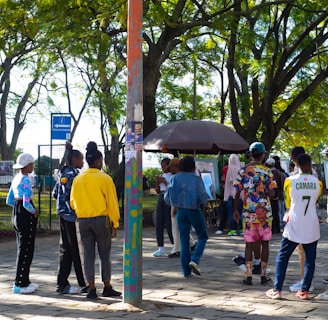 A group of people gathers in a park under leafy trees. They are interacting near a kiosk or stand surrounded by greenery and paved ground. Some individuals are wearing colorful clothing, and one person is wearing a number 7 sports jersey. There's a mixture of standing and walking activities happening in the scene.