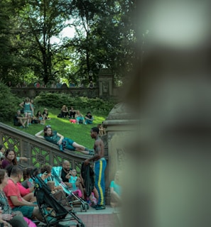 A lively outdoor scene in a park featuring several people sitting and interacting on stone steps and benches. A man in athletic wear stands by a group, engaging in conversation. The lush greenery of trees provides a natural canopy in the background.
