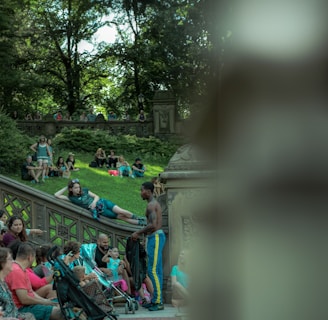 A lively outdoor scene in a park featuring several people sitting and interacting on stone steps and benches. A man in athletic wear stands by a group, engaging in conversation. The lush greenery of trees provides a natural canopy in the background.